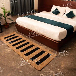 May include: A black and beige jute rug with a striped pattern. The rug is placed in front of a bed with white linen and a teal runner. The text "Tulsi India Home Decor Rugs" is visible throughout the image.