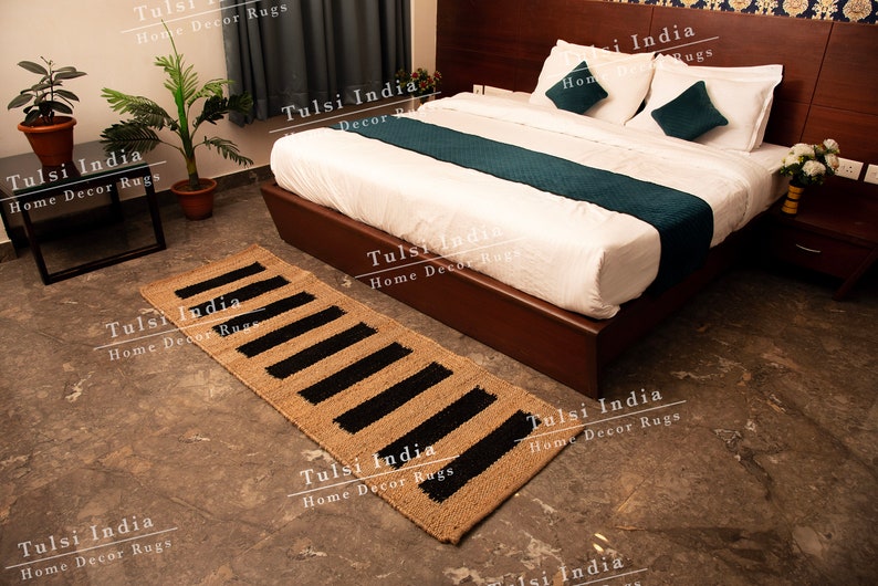 May include: A black and beige jute rug with a striped pattern. The rug is placed in front of a bed with white linen and a teal runner. The text "Tulsi India Home Decor Rugs" is visible throughout the image.