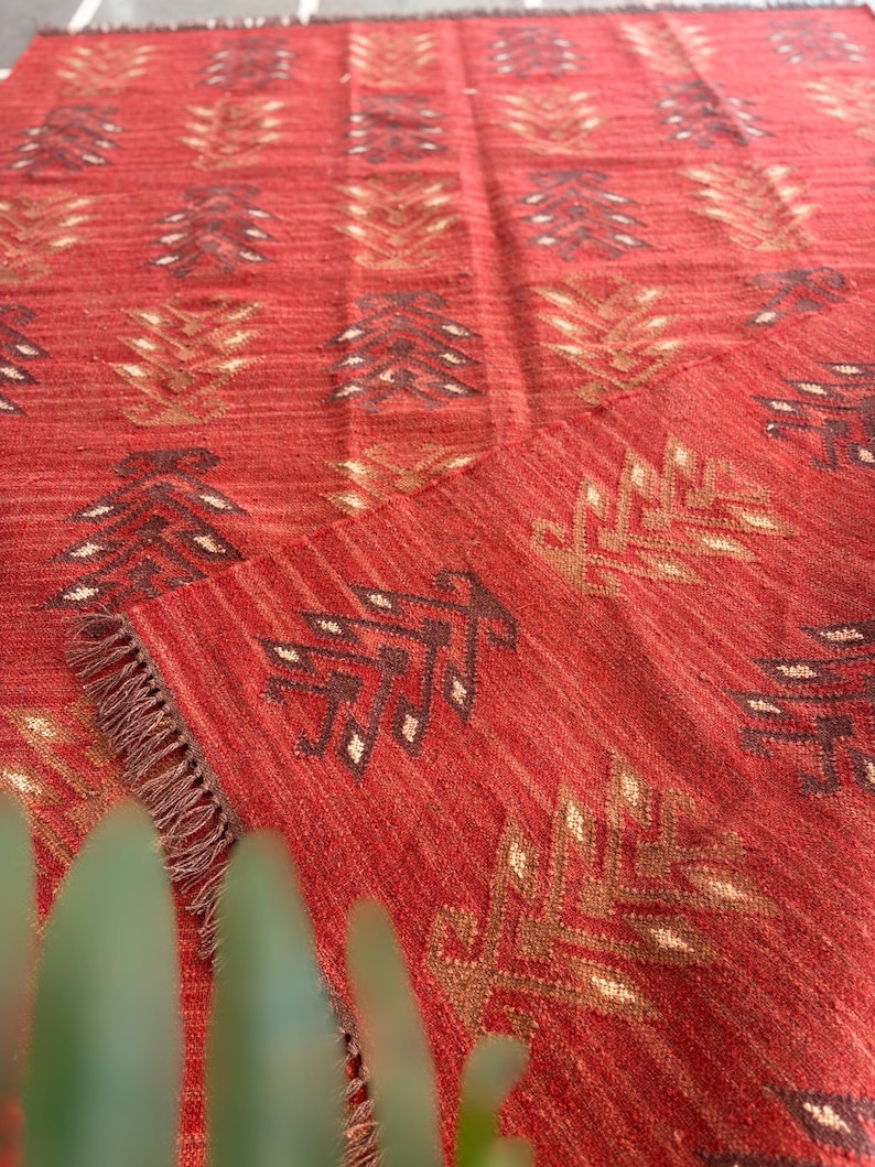 May include: Red area rug with a geometric pattern of brown and gold leaves. The rug has a fringe border.