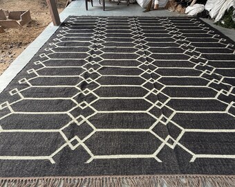 May include: A large, rectangular rug with a geometric pattern. The rug features a dark gray background with a repeating white geometric design. The rug has a fringe along the bottom edge. This rug is suitable for a living room or bedroom.