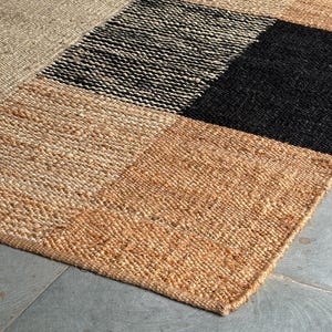 Op de afbeelding: Een rechthoekig jute vloerkleed met een patchwork ontwerp. Het kleed heeft vierkanten in beige, zwart en verschillende bruintinten. De geweven textuur is zichtbaar, wat een natuurlijk en gestructureerd uiterlijk creëert. Het kleed ligt op een grijze tegelvloer.