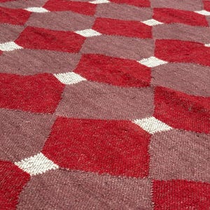 May include: Close-up of a woven rug with a geometric pattern. The rug features a repeating design of red and mauve diamond shapes, separated by white diamond accents. The texture of the rug is visible.
