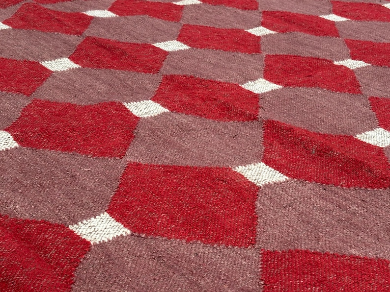 May include: Close-up of a woven rug with a geometric pattern. The rug features a repeating design of red and mauve diamond shapes, separated by white diamond accents. The texture of the rug is visible.