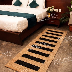May include: A natural fibre rug with black stripes. The rug is made of jute and is perfect for adding a touch of natural beauty to any room. The rug is shown in a bedroom setting, placed in front of a bed with white bedding and teal throw pillows.