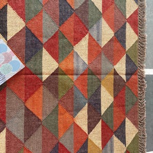 May include: A geometric patterned rug with a fringe border. The rug is made of woven jute and features a variety of colours, including red, orange, brown, green, and beige.