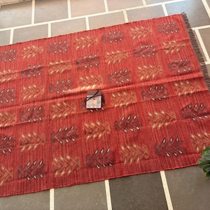 May include: A red and white patterned rug with fringed edges. The rug has a geometric design with a repeating pattern of stylized leaves.