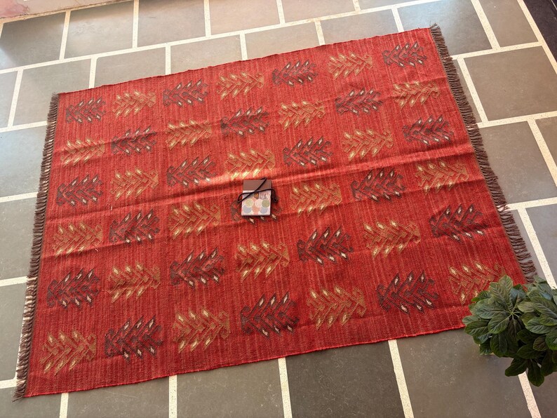 May include: A red and white patterned rug with fringed edges. The rug has a geometric design with a repeating pattern of stylized leaves.