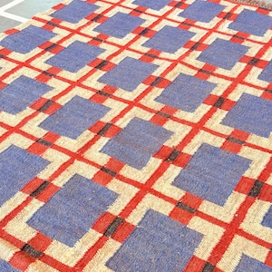 May include: A blue and red geometric patterned rug with a woven texture. The rug has a square design with a grid pattern.