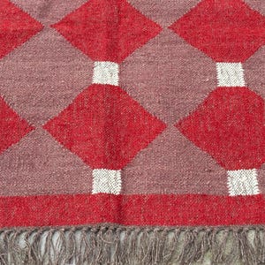 May include: A close-up of a woven rug with a geometric pattern. The rug features a repeating design of red diamonds, separated by mauve squares and white rectangles. The rug has a fringe along the bottom edge.