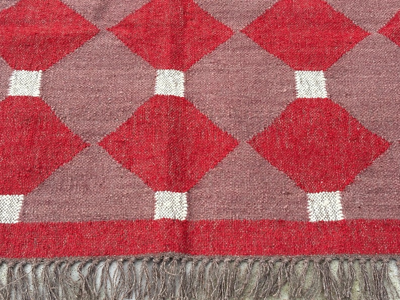 May include: A close-up of a woven rug with a geometric pattern. The rug features a repeating design of red diamonds, separated by mauve squares and white rectangles. The rug has a fringe along the bottom edge.
