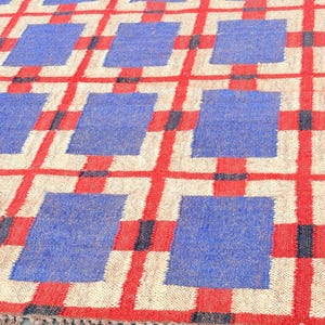 May include: A woven rug with a geometric pattern of red, blue, and white squares. The rug has a fringe border.