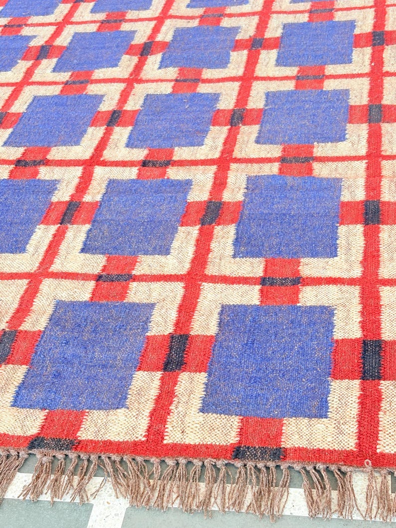 May include: A woven rug with a geometric pattern of red, blue, and white squares. The rug has a fringe border.