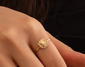 May include: A gold ring with a lion head design. The ring is worn on a finger.
