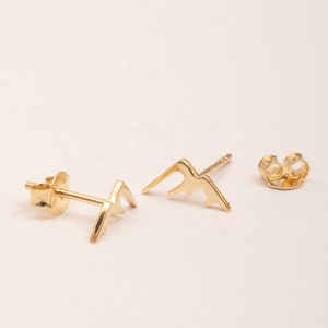 May include: A pair of gold mountain-shaped stud earrings. The earrings are small and delicate, and they have a simple, minimalist design.