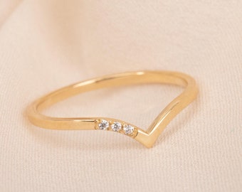 May include: A gold-colored V-shaped ring with three small, clear gemstones set along the band. The ring is displayed on a cream-colored fabric, highlighting its delicate design and sparkle. This piece of jewelry is suitable for everyday wear or special occasions.
