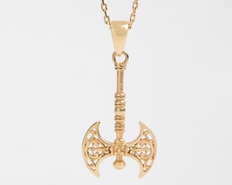 May include: A gold-colored pendant necklace featuring a detailed axe charm. The axe charm has an ornate, openwork design on the double-bladed head. The necklace chain is also gold-colored and hangs against a white background.
