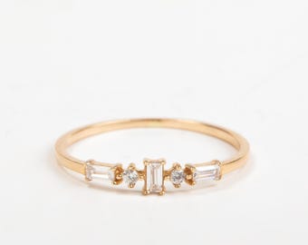 May include: A delicate gold ring featuring a linear arrangement of clear, rectangular and round-cut gemstones. The ring's band is thin and polished, with the gemstones set in a row across the top. The ring is set against a plain white background.