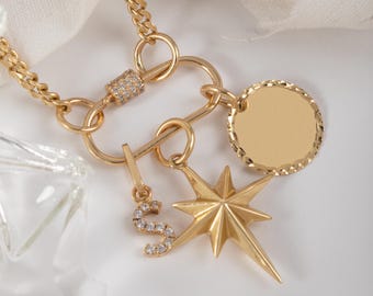 May include: A gold-colored necklace with multiple charms. The necklace features a curb chain, a clasp with embedded crystals, a starburst charm, a circular charm, and a charm with the letter "S" encrusted with crystals. The background is white.