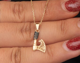 May include: A gold-colored pendant necklace featuring a small axe charm. The axe head has a Celtic knot design, and the handle is wrapped with black detailing. The necklace chain is also gold-colored.