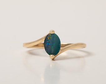 May include: A gold ring featuring a central oval-shaped dark blue opal gemstone with iridescent green and orange flecks. The band is smooth and polished, with a bypass design that secures the stone at the top and bottom with small gold prongs.