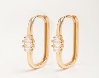 May include: A pair of gold-colored hoop earrings. Each earring has a rectangular shape with a cluster of clear, rectangular-cut stones in the center. The earrings are displayed against a white background.