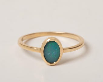 May include: A gold ring featuring a central oval-shaped dark green opal gemstone with subtle blue and green iridescent flecks. The band is thin and smooth, crafted from a polished yellow gold metal, creating a simple and elegant jewelry piece.