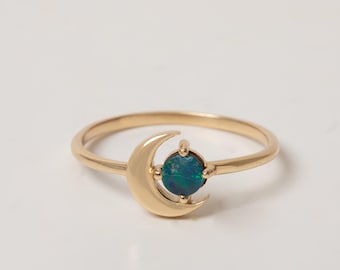 May include: A gold ring featuring a crescent moon design and a small, round, dark blue-green opal gemstone set in the center. The band is thin and polished, with the moon shape cradling the circular stone in a prong setting on the top of the ring.