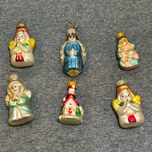 May include: Six vintage glass Christmas ornaments featuring angels and a church. The ornaments are all white with gold and silver accents. The angels are wearing blue, green, and white robes. The church is red and white with a green tree.