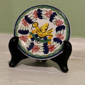 May include: A small, white ceramic plate with a yellow bird design. The bird is surrounded by pink and blue flowers. The plate is on a black stand.