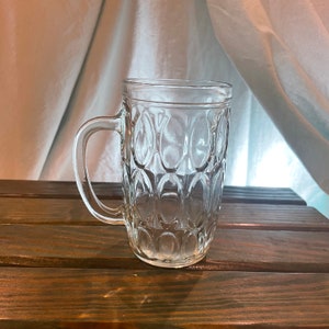 May include: A clear glass mug with a textured, bubble-like design. The mug has a handle and is sitting on a wooden surface.