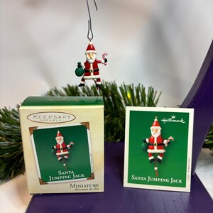 May include: A Hallmark Keepsake Christmas ornament featuring a miniature Santa Jumping Jack. The ornament is red, white, and black, with a green bag and candy cane. The box and card also feature the Santa Jumping Jack design.