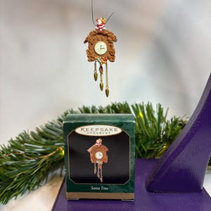 May include: A brown cuckoo clock Christmas ornament with a Santa Claus figure on top. The clock has a white clock face and gold-colored pendulum and weights. The ornament is suspended above a green box with the words "Keepsake Ornament" and "Santa Time".