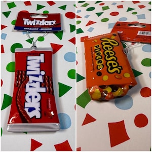 May include: Two candy-themed ornaments featuring a red Twizzlers package with white text and a silver hanging chain, and an orange Reese's Pieces package with yellow and black text, both attached to cardboard backing cards with hanging holes.