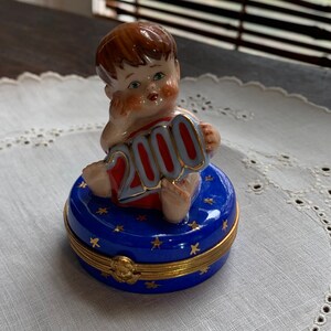 May include: A porcelain figurine of a child sitting on a blue and gold trinket box. The child is holding a red and gold sign that says "2000".