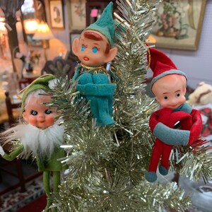 May include: Three vintage felt Christmas elf ornaments hanging on a silver tinsel tree. The elves are wearing green, blue, and red outfits with white fur trim.