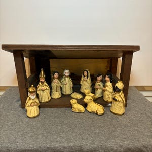 May include: A vintage nativity scene set with a wooden stable and 12 ceramic figurines, including Mary, Joseph, baby Jesus, shepherds, and animals. The figurines are painted in a cream color with gold accents.