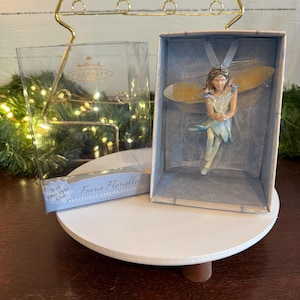 May include: A blue and yellow fairy figurine ornament with fiber optic glow. The ornament is in a clear plastic box with the text "Faerie Floriella" and "Hallmark Keepsake Collection".