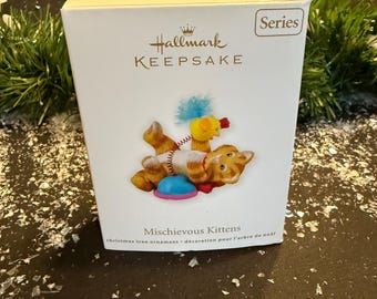 Könnte beinhalten: Eine Hallmark Keepsake-Box mit einem Weihnachtsbaumschmuck. Der Schmuck zeigt zwei orangefarbene Tabby-Kätzchen, die mit einem Spielzeug spielen. Auf der Box stehen die Worte "Mischievous Kittens" und "christmas tree ornament - décoration pour l'arbre de noël".