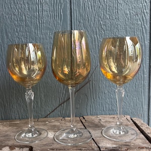 May include: Three iridescent gold wine glasses with clear stems and bases. The glasses have a rounded bowl shape and are displayed on a wooden surface against a blue background. The stems have a decorative design.