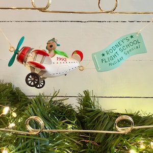 May include: A white and red airplane ornament with a green propeller and a brown bear wearing a green hat and goggles. The airplane is pulling a banner that says "Rodney's Flight School Special Holiday".