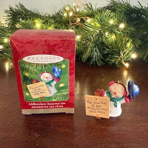 May include: A red box with a white snowman ornament inside. The ornament is holding a sign that says "Tip your hat to the millennium 2000-2001". The box has the words "Keepsake Ornament" on it.
