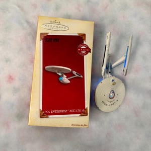 May include: A Hallmark Keepsake Ornament of the U.S.S. Enterprise NCC-1701-A starship. The packaging is a rectangular box with a red center panel, white borders, and the text Star Trek and U.S.S. Enterprise NCC-1701-A. The model is white and gray.