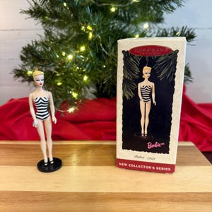 May include: A Barbie doll ornament in a black and white striped swimsuit stands next to its original box. The box features an illustration of the doll and the text "Debut - 1959" and "New Collector's Series". A Christmas tree is in the background.