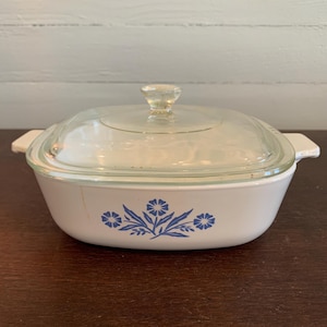 May include: White casserole dish with a clear glass lid. The dish features a blue floral design and side handles. The lid has a clear knob. A vintage kitchenware item.