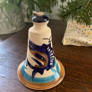 May include: A white ceramic bell with a blue and brown marlin design. The bell has a blue top and a brown rim.