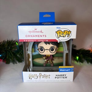 May include: A Harry Potter Christmas tree ornament in a display box. The ornament is a Funko Pop! figure of Harry Potter, wearing glasses and holding a wand. The box has the Hallmark and Walmart logos.