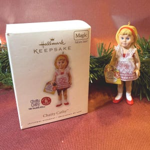 May include: A Hallmark Keepsake Christmas ornament featuring a doll named Chatty Cathy. The doll has blonde hair, blue eyes, and wears a red and white dress. The box next to the doll has the text "Chatty Cathy" and "Magic Features Sound."
