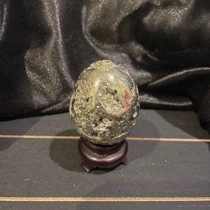 May include: A pyrite egg-shaped stone, with a metallic, reflective surface, sits on a small, dark wooden stand. The stone has a rough, textured appearance with small cavities. The background is a dark, draped fabric.