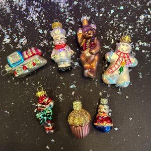 May include: A collection of Christmas ornaments in various shapes, including a train, chef, bear, gingerbread man, candy cane, cupcake, and snowman. The ornaments are made of glass and feature vibrant colors and glitter accents. Each ornament has a gold-colored metal cap for hanging.