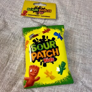 May include: A Sour Patch Kids ornament shaped like a yellow candy bag with green accents. The front features the Sour Patch Kids logo, the text Sour Then Sweet, and various colorful gummy candy figures. It includes a yellow cardboard header tag.